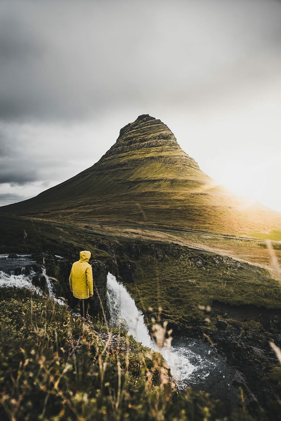 person standing on cliff near waterfalls during daytime, person in yellow hooded jacket standing with brown mountain background HD wallpaper
