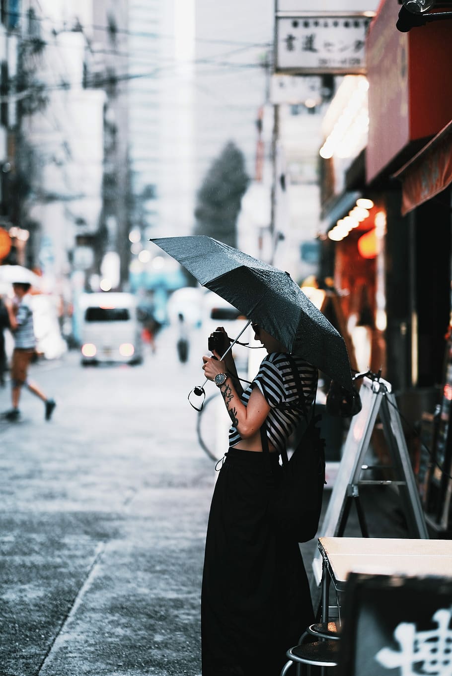 Japan 2017, woman holding umbrella and point-and-shoot camera while standing beside table and signage in street during daytime