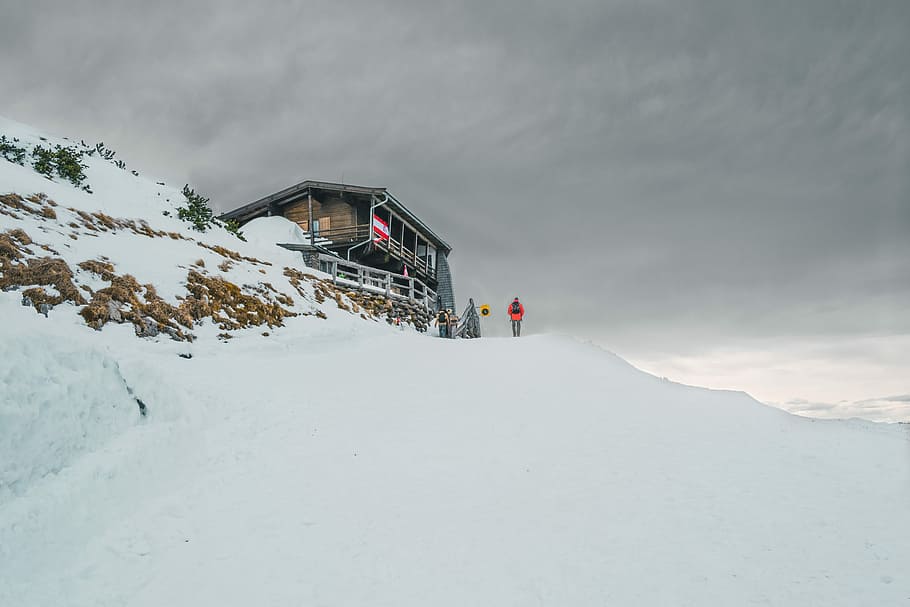 person standing on glacier hill near house under white cloudy skies during winter, group of man standing near brown wooden house HD wallpaper