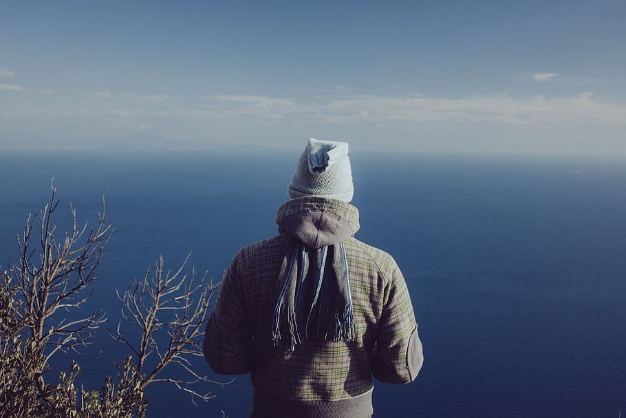 man wearing hooded jacket and gray cap overlooking large blue body of water during daytime HD wallpaper