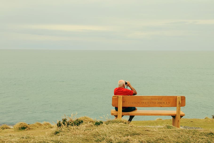 man wearing red t-shirt sitting on brown wooden bench facing on body of water, person holding camera sitting on bench facing ocean during daytime HD wallpaper