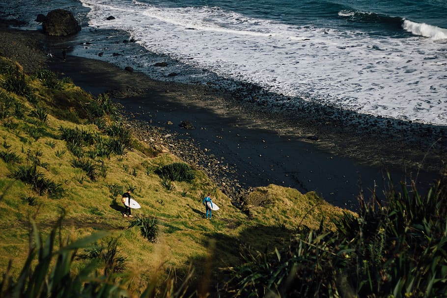 two person holding surfboard on seashore, aerial photography of two person walking down hill towards beach shore while carrying surfboards HD wallpaper