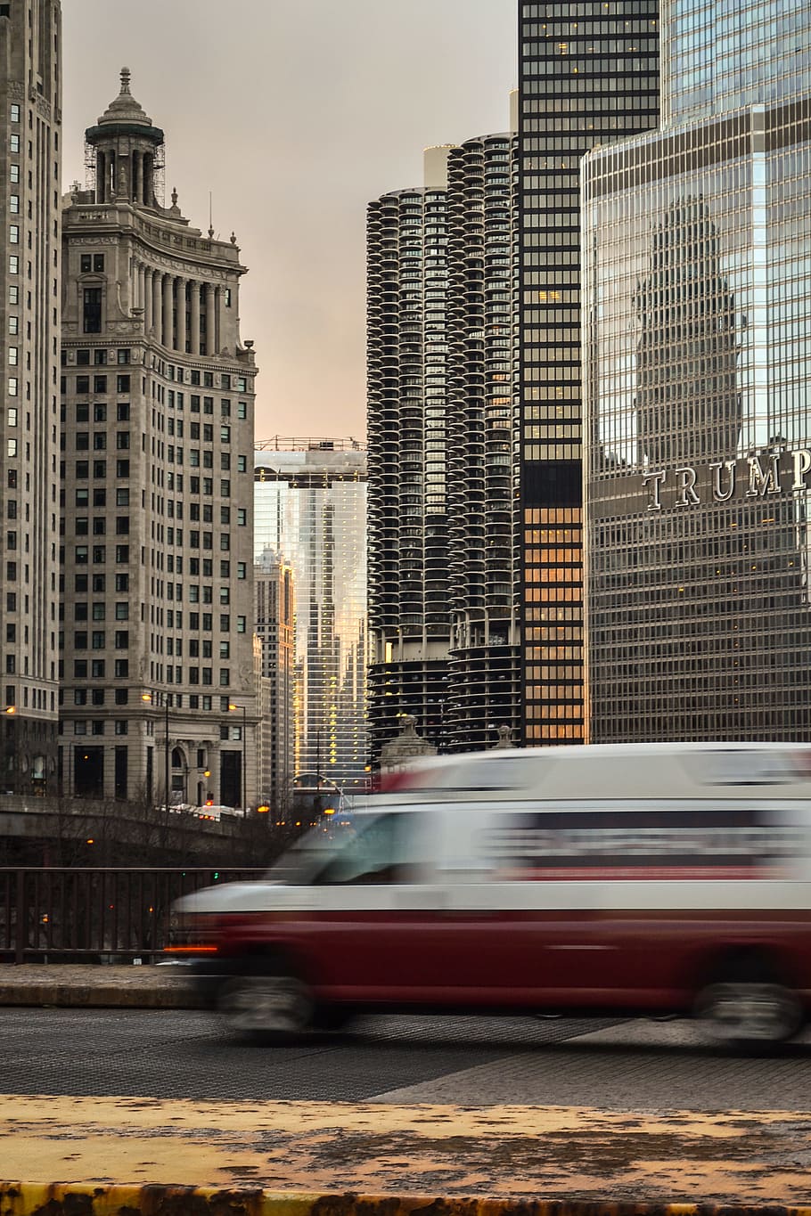A blurry car on a bridge in a city with skyscrapers at the back, red and white van on road near glass building at daytime