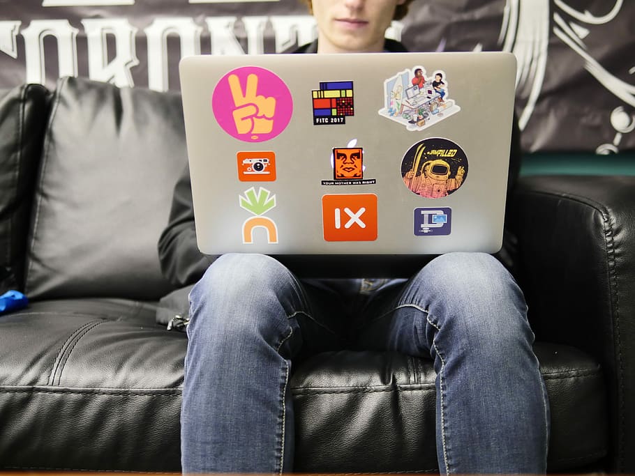 man using silver MacBook sitting on black leather sofa, person wearing blue denim jeans sitting using gray laptop