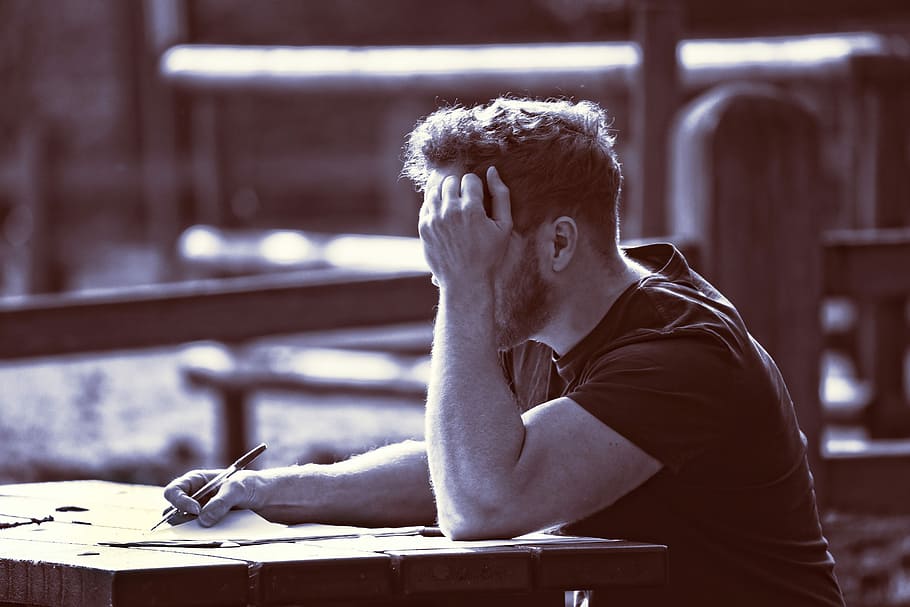 man sitting at table with left hand near face and right hand holding paper and pen HD wallpaper