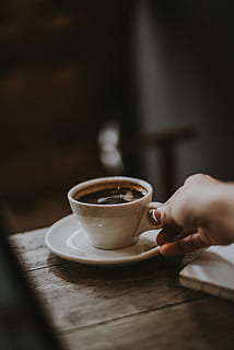 person holding white ceramic cup with liquid, selective focus photography of person holding ceramic mug with black liquid on brown table HD wallpaper