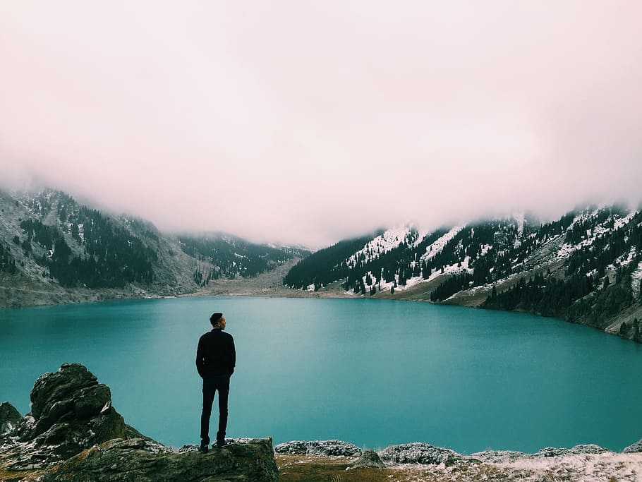 man standing on mountain looking at lake under white clouds at daytime, man standing on gray cliff in front of lake