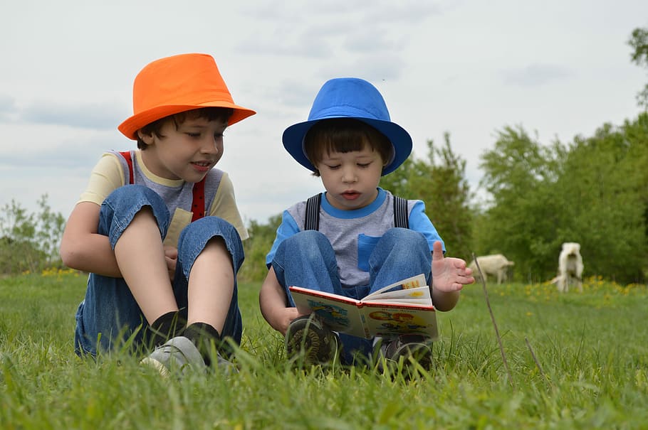 kid reading a book beside boy while sitting on grass field outdoor