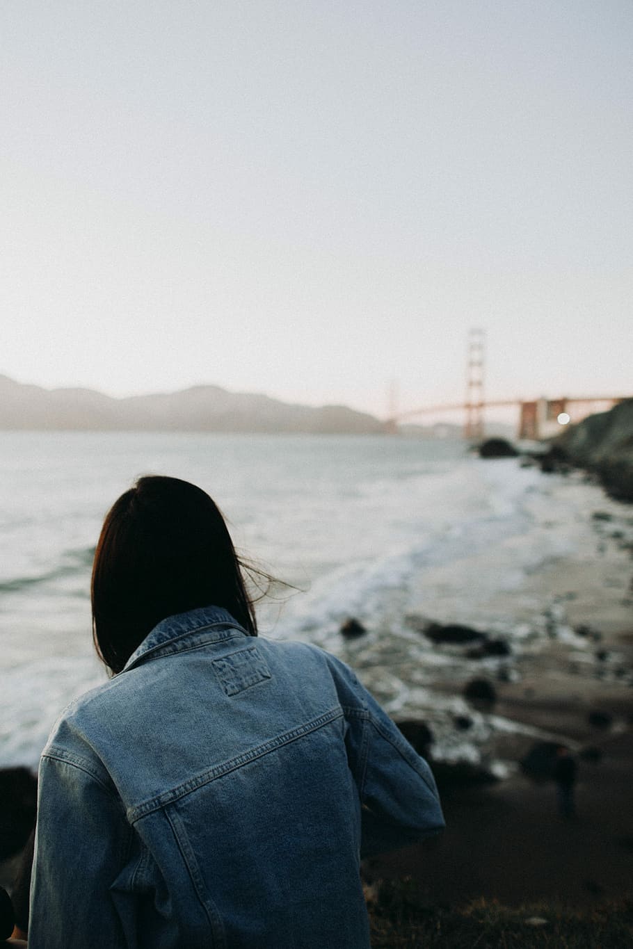 person sitting near sea overlooking red bridge during daytime, woman wearing blue denim jacket sitting on rock near seashore HD wallpaper