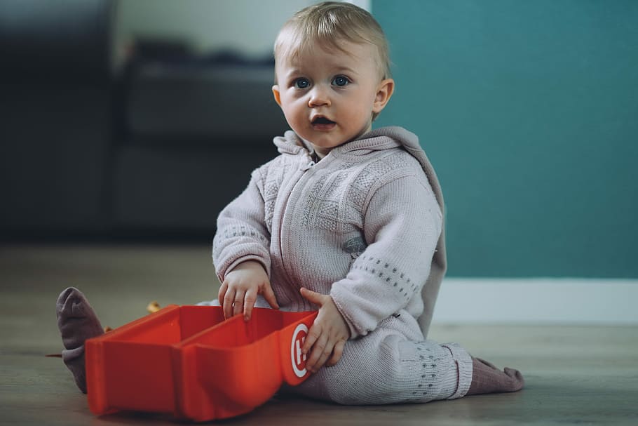 toddler sitting on ground while holding red plastic case, baby wearing gray jacket holding a red plastic toy while sitting on floor