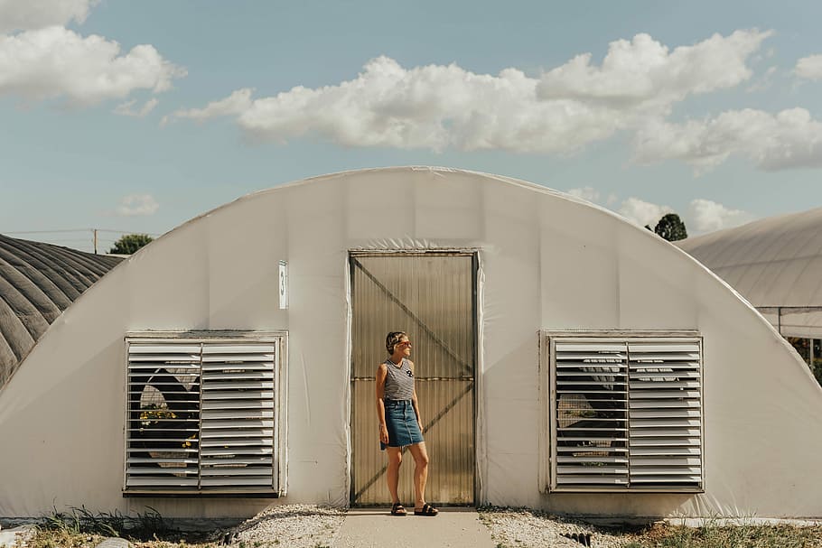 girl in gray tank top behind white shed, woman standing near white and gray half-moon wall outside during daytime photography