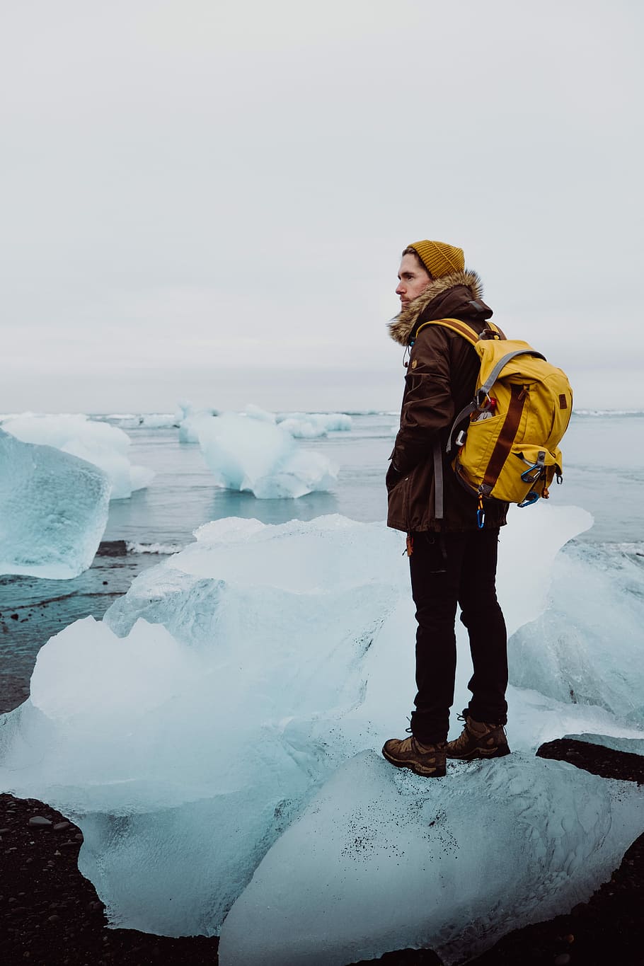 man standing on ice rack under gray sky, man standing on iceberg in in front of body of water HD wallpaper
