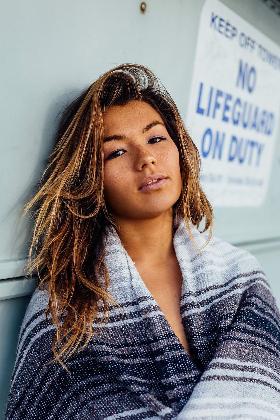 woman with black, white, and grey blanket leaning on wall with no lifeguard on duty signage, woman in white, gray, and black top