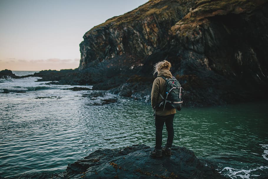 photo of person in brown hoodie, green backpack, black pants, and brown shoes standing on rock near ocean, person standing on boulder HD wallpaper