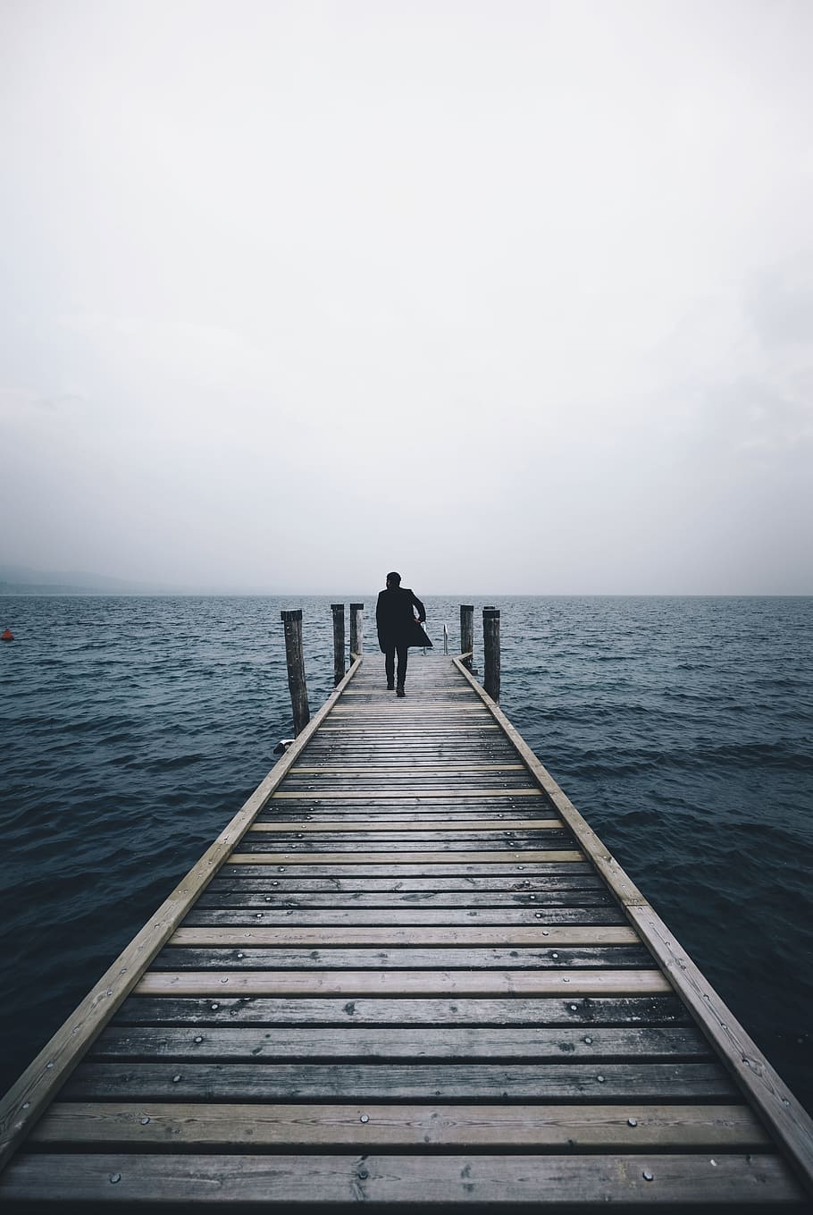 man walking on dock surrounded by ocean during daytime, man in black suit walks on brown wooden dock in middle of sea under cloudy sky during daytime, HD wallpaper