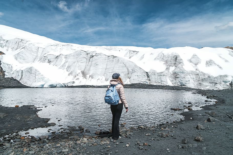 person standing on body of water near snowy mountain, woman in white jacket and black pants standing beside lake HD wallpaper