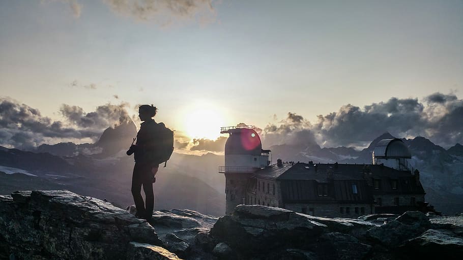 silhouette of woman with backpack on rock near brown building at sunset, silhouette of person standing on rock formation during daytime HD wallpaper