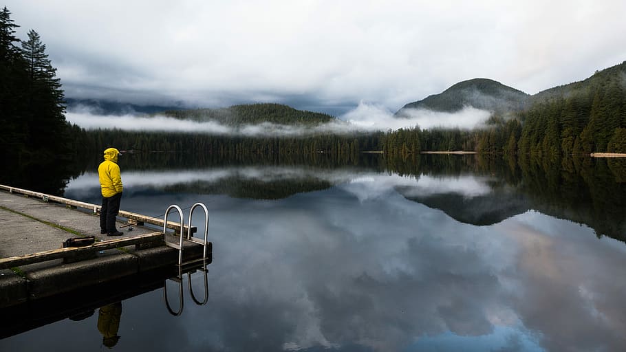 person standing near edge near body of water, man standing on corner of dock facing body of water with trees and mountain in background HD wallpaper
