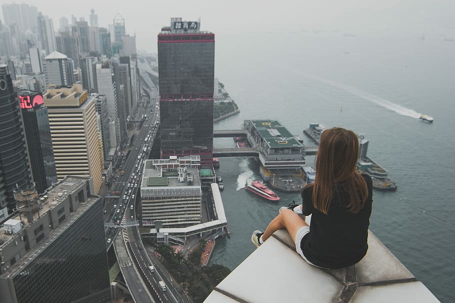 woman sitting on top of building overlooking on city by the bay during daytime, woman sitting on building while looking at cityscape near body of water