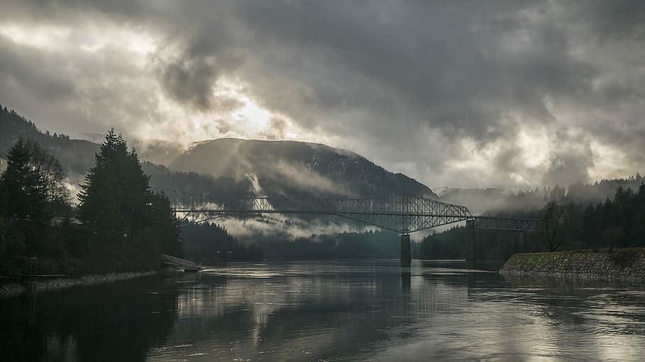 green trees by the lake near mountain during daytime, photo of grey metal bridge under cloudy sky HD wallpaper