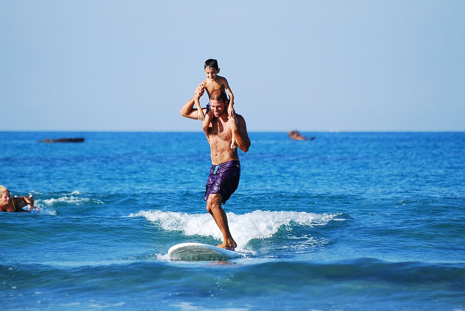 man surfing carrying boy, man and son carrying shoulder riding surfboard during daytime