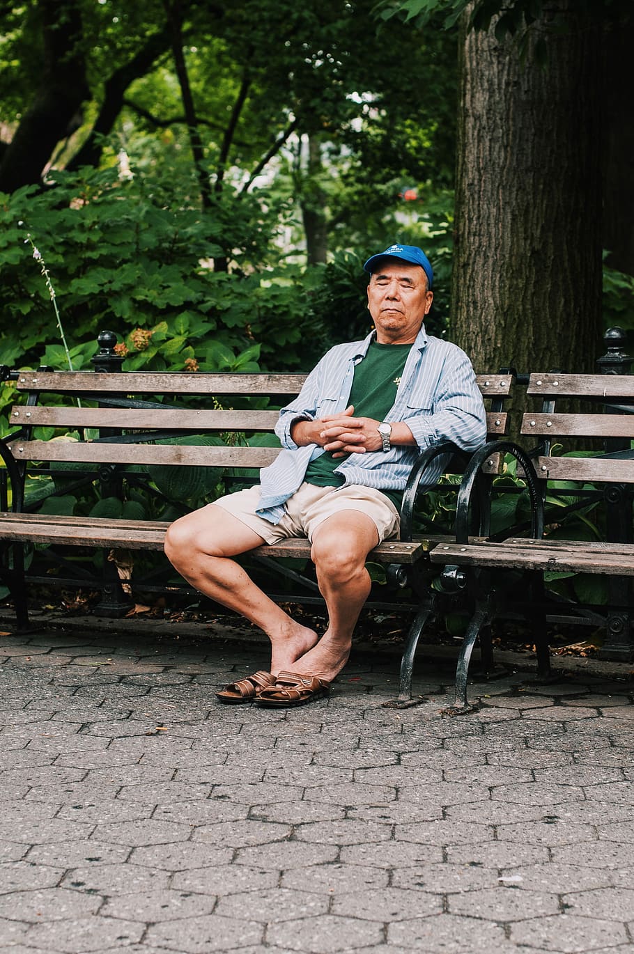 man sitting on bench closing his eyes near trees, sitting man wearing blue striped sports shirt and khaki shorts on bench at park