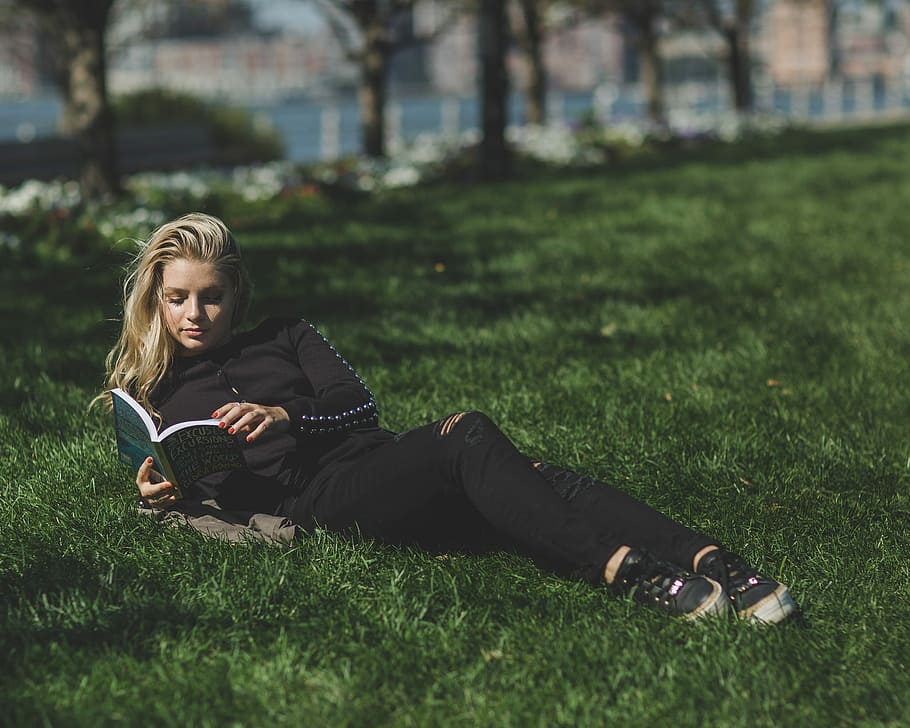 woman reading book leaning on green grass field, woman in black long-sleeved shirt and black pants lying on green grass while reading during daytime