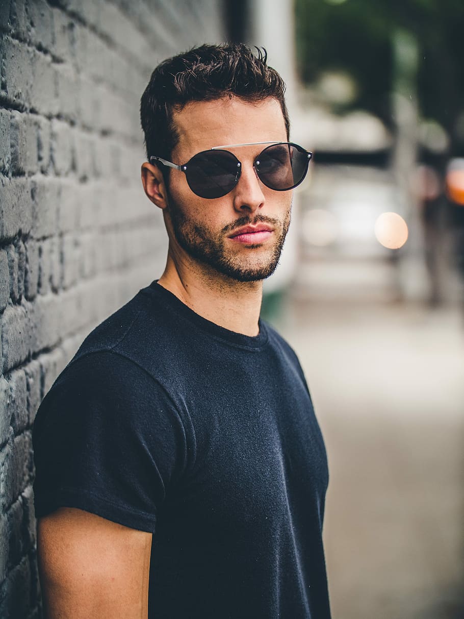 man standing beside building wall, man wearing black crew-neck t-shirt and gray framed sunglasses