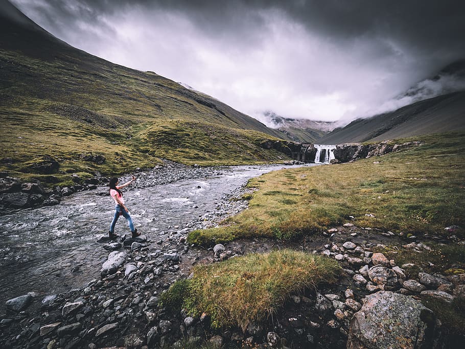 woman in blue jeans crossing river, woman wearing pink shirt and blue denim jeans standing on river between green grass field under gray and white cloudy sky HD wallpaper