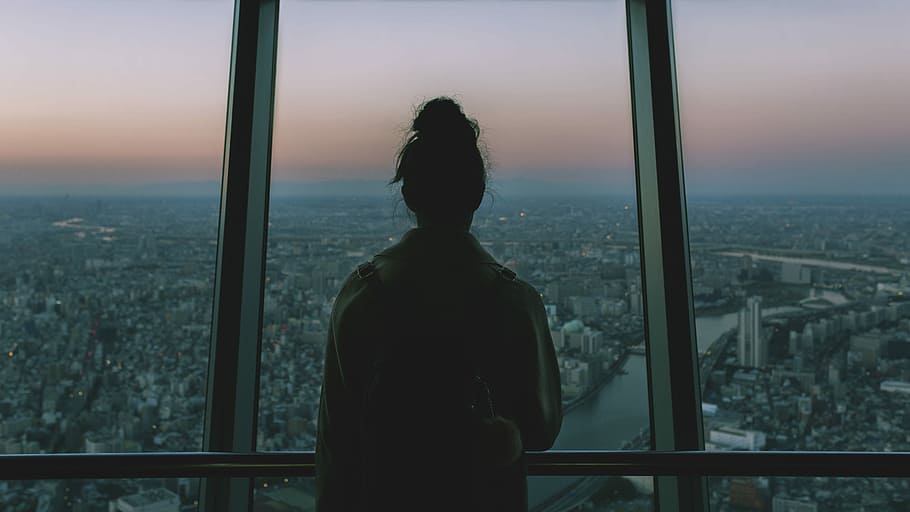 silhouette of person standing inside the building facing the glass mirror watching the city, woman looking through window in city photography