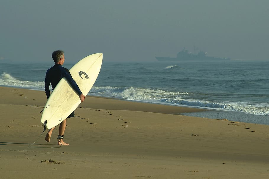 man carrying white surfboard standing on shore during daytime HD wallpaper
