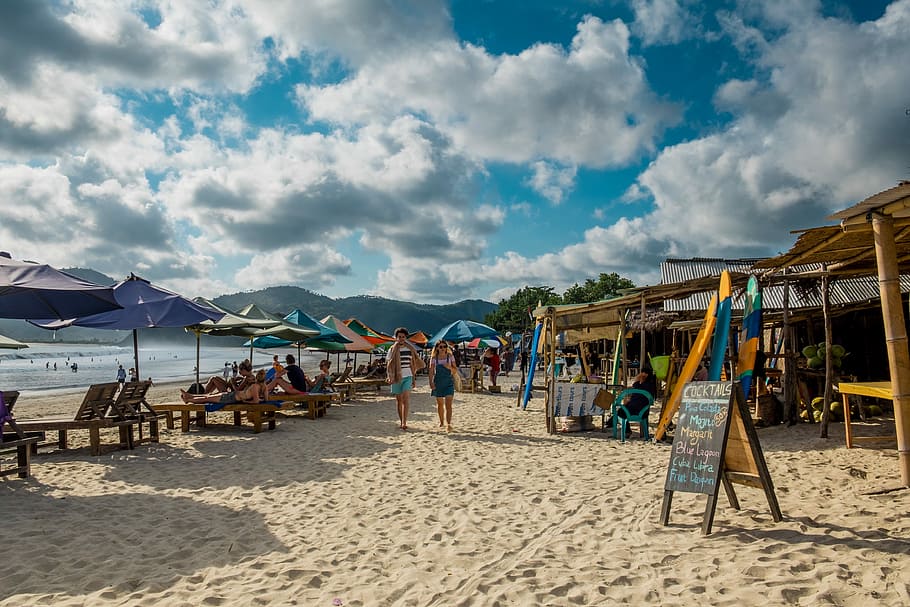 two person walking on beach with assorted umbrellas and nipa huts, two person walking across beach stalls near beach under white cloudy skies HD wallpaper