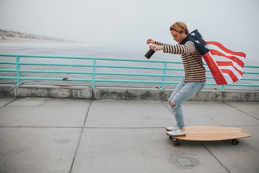 man wearing US flag cape while riding on longboard near body of water during daytime, man in brown sweater holding america flag on brown longboard during daytime HD wallpaper