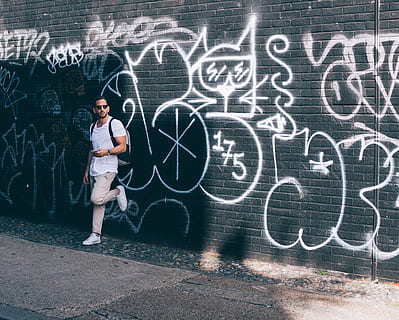 man wearing white crew-neck T-shirt leaning on black wall with white graffiti, man leaning on graffiti wall HD wallpaper