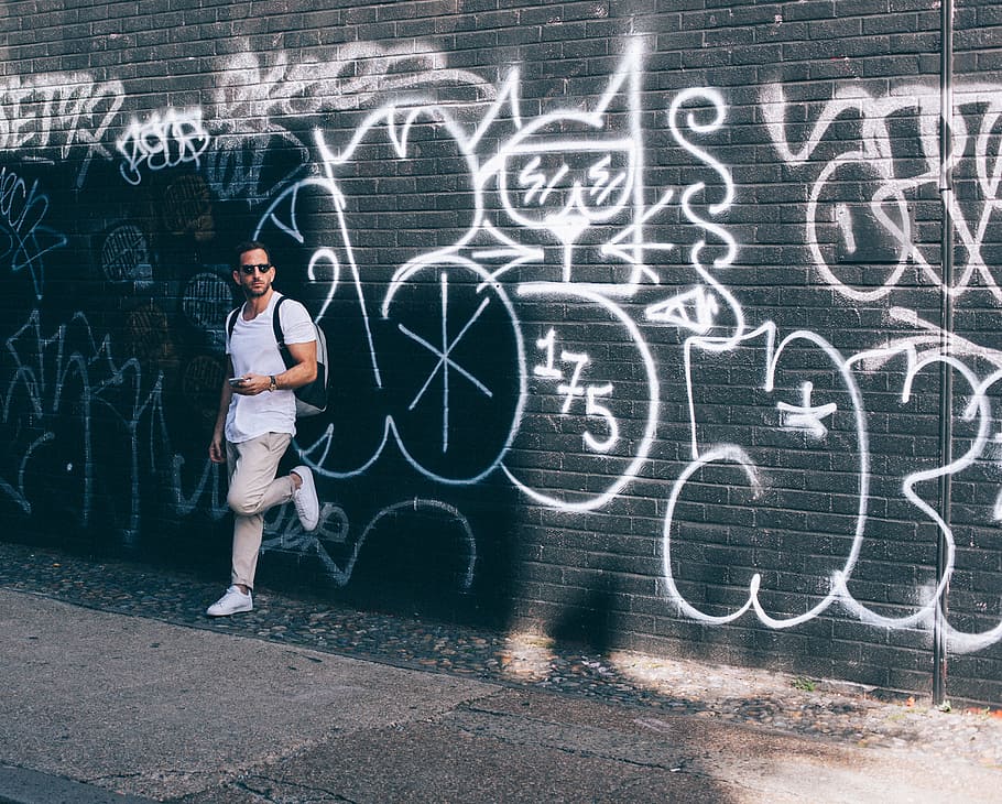 man wearing white crew-neck T-shirt leaning on black wall with white graffiti, man leaning on graffiti wall HD wallpaper