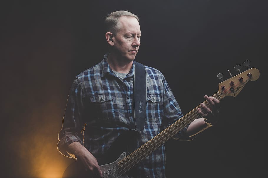 man playing electric guitar in dark room, focus photo of man in gray and blue plaid button-up long-sleeved shirt playing white and black 4-string bass guitar HD wallpaper