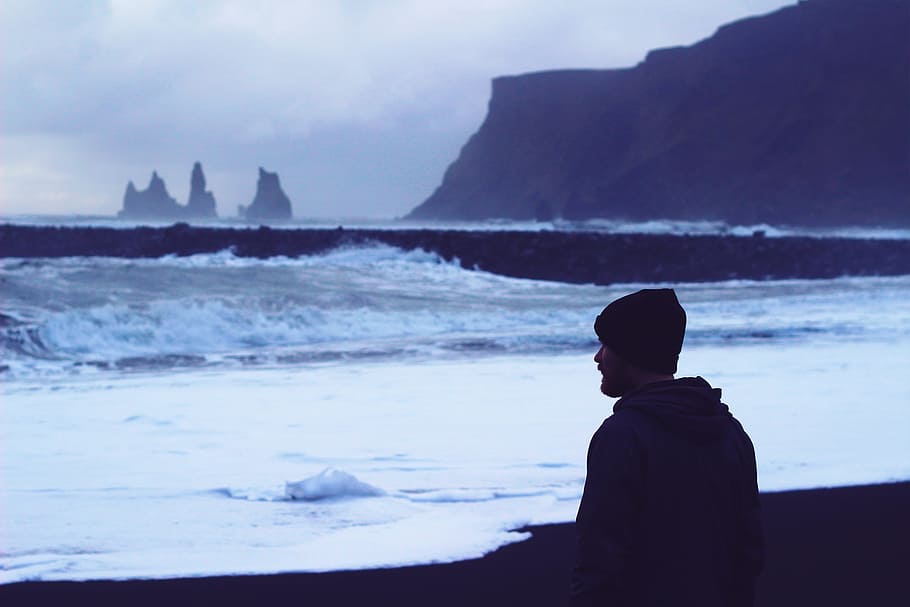man standing on seashore looking at water waves, man wearing knit cap and jacket near body of water