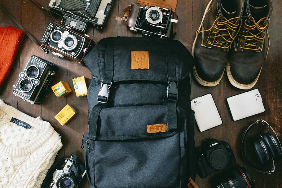 high-angle photo of black bag beside cameras, portable HDD and black leather lace-up boots, black backpack and pair of black sneakers on brown floor HD wallpaper