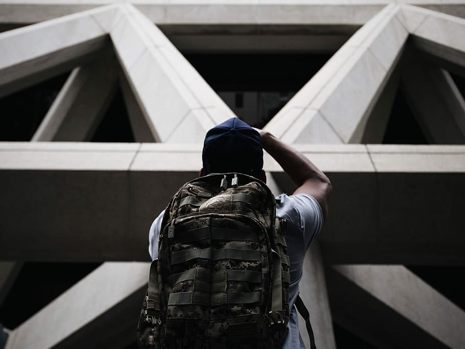 person wearing shirt and hiking bag looking in front of grey concrete structure while standing during daytime, man in white shirt with backpack and blue hat looking up in concrete building during daytime HD wallpaper