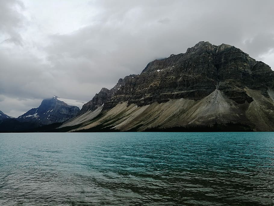 black and brown rocky mountain beside body of water under cloudy sky, mountain and body of water under black sky HD wallpaper
