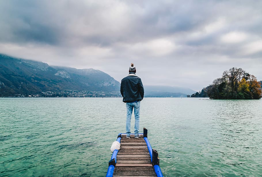 man standing on edge of dock facing mountains, man standing on dock facing body of water HD wallpaper
