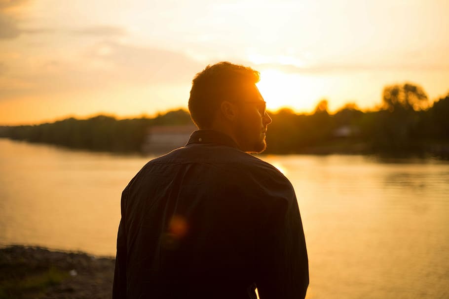 man wearing sunglasses in front of calm body of water during golden hour HD wallpaper