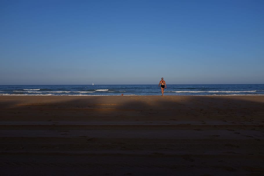 woman in black one-piece swimsuit facing blue wavy ocean under clear blue sky, person walking on shore during daytime