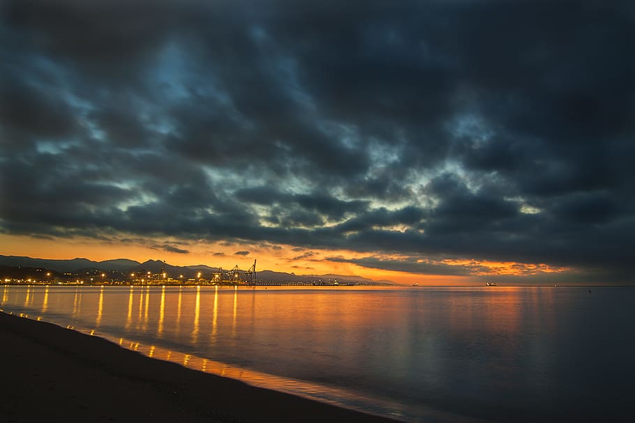 lighted city under stormy sky during daytime, calm body of water against orange sky HD wallpaper