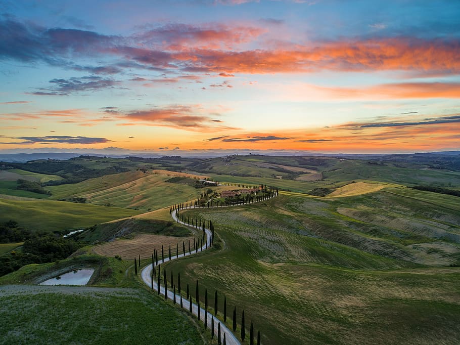 road between green grass field near mountains under blue and brown sky at golden hour, top view photo of asphalt road between grass field HD wallpaper