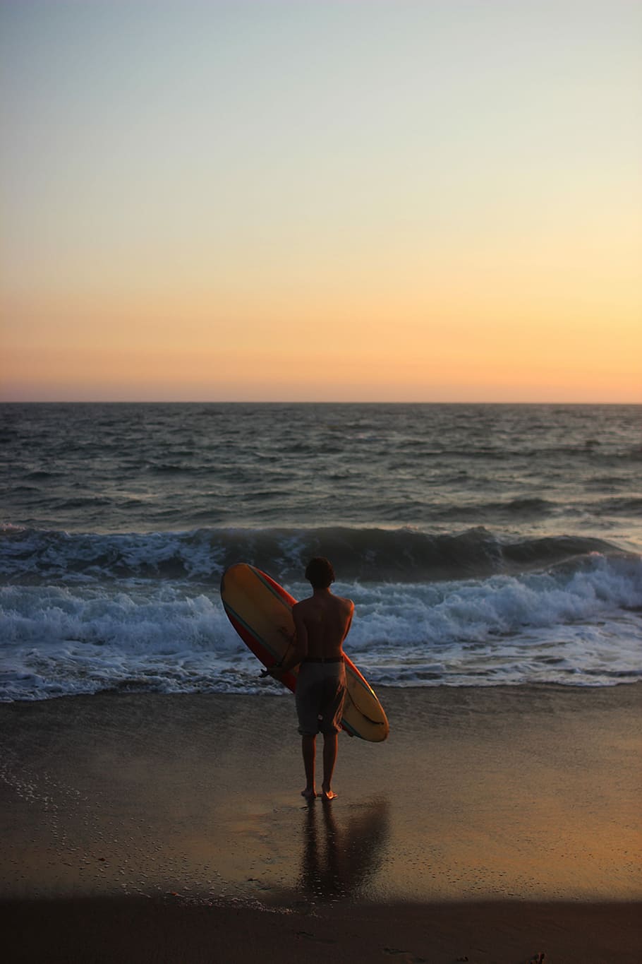 man standing on shore during daytime while carrying beige surfboard, person holding surfboard in front of body of water HD wallpaper