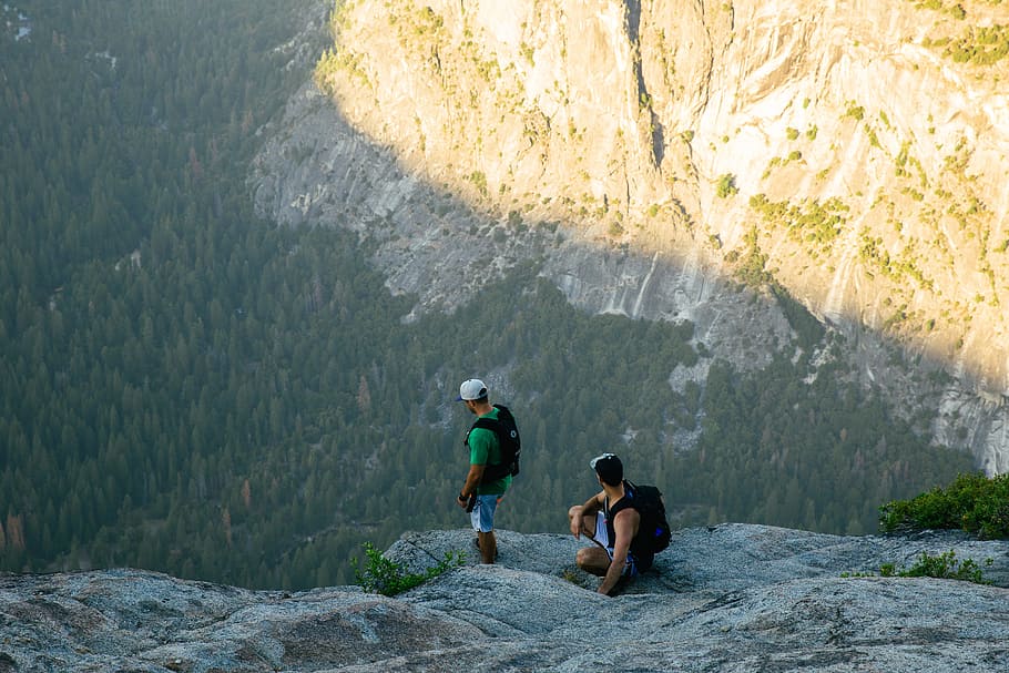 two men standing on rocky mountain cliff, two men standing on rock ledge facing on cliff at daytime HD wallpaper