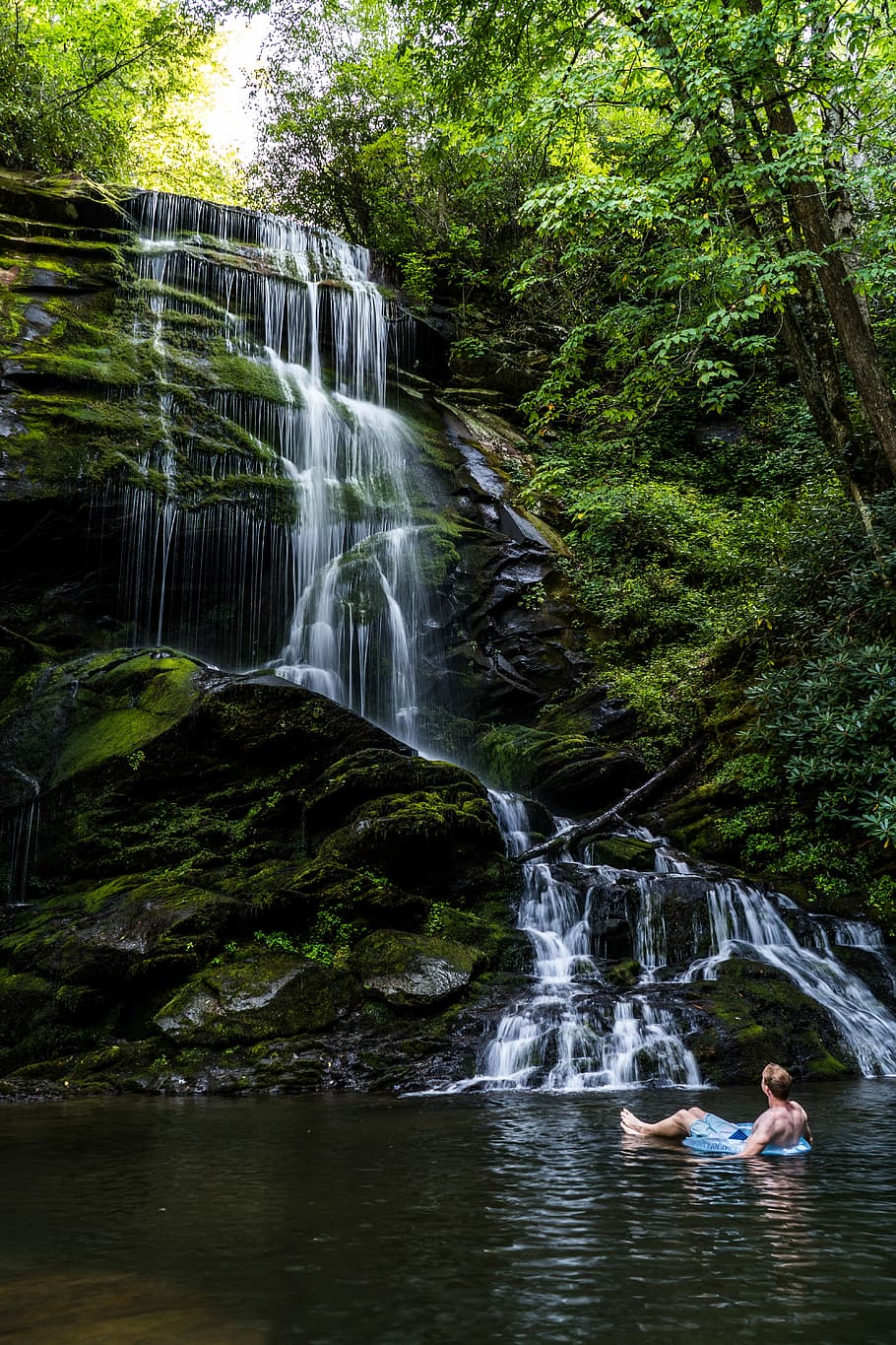 man riding blue inflatable ring in body of water while looking up watching the waterfalls at daytime, man on inflatable floater floating near waterfalls HD wallpaper