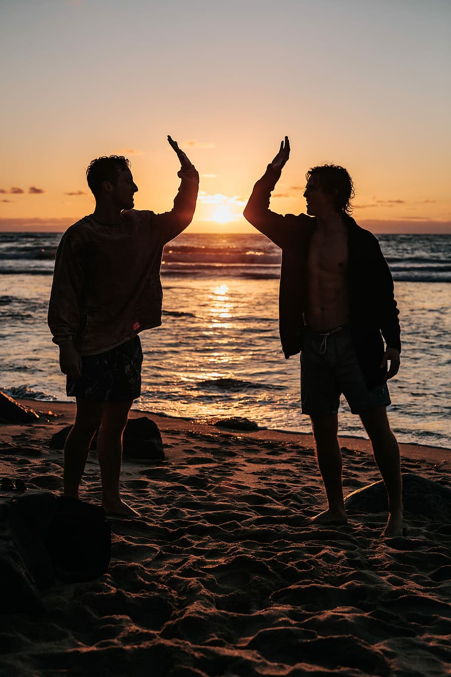 silhouette of two mens near seashore about to high five during sunset, silhouette of two men doing high five standing on sandy beach during sunset