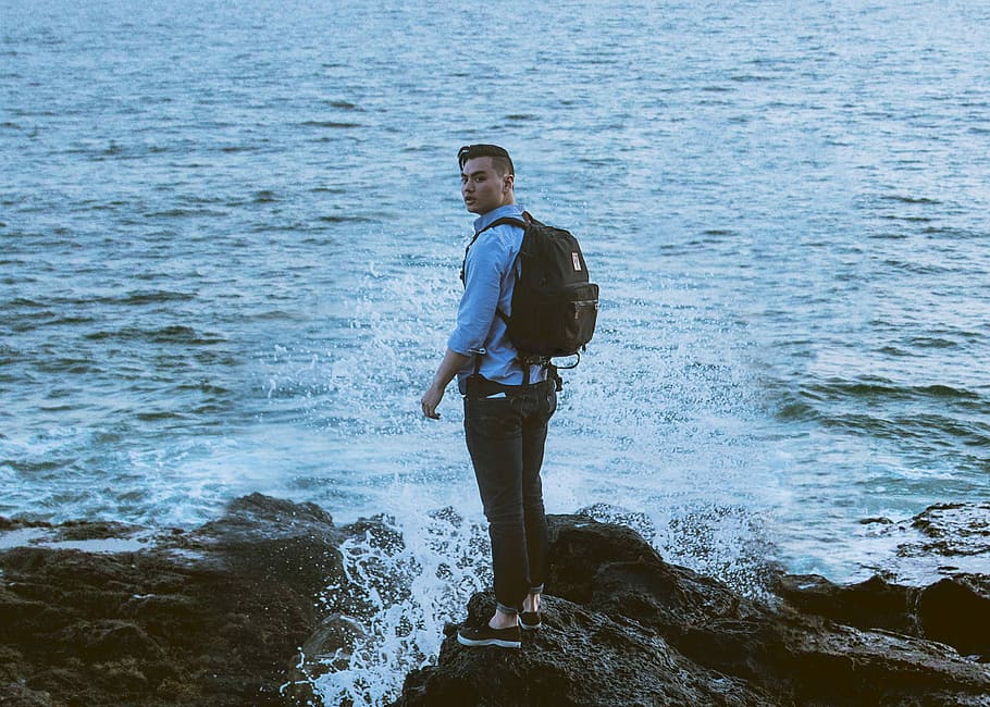 man standing on gray rock formation near body of water wearing backpack, man standing near body of water HD wallpaper